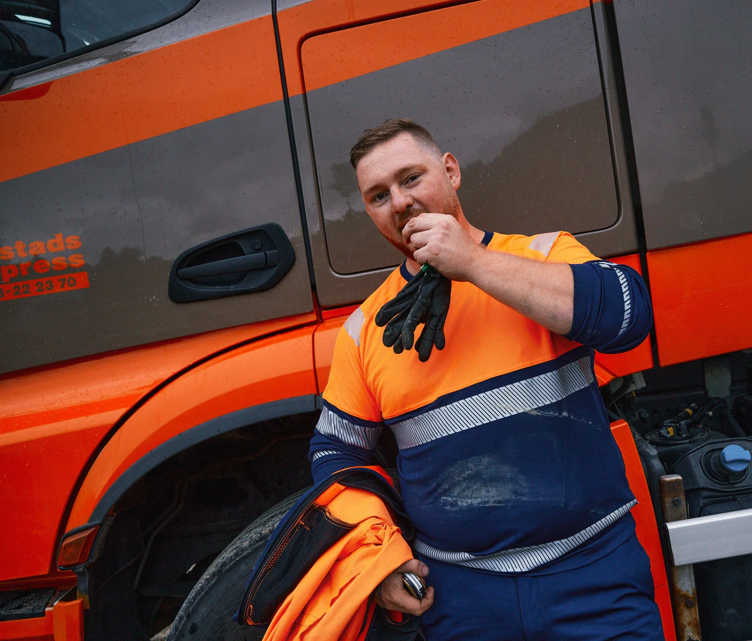 Truck driver driver wearing a orange/navy high-visibility jacket and work trousers from Fristads.