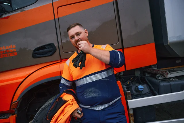 Truck driver driver wearing a orange/navy high-visibility jacket and work trousers from Fristads.